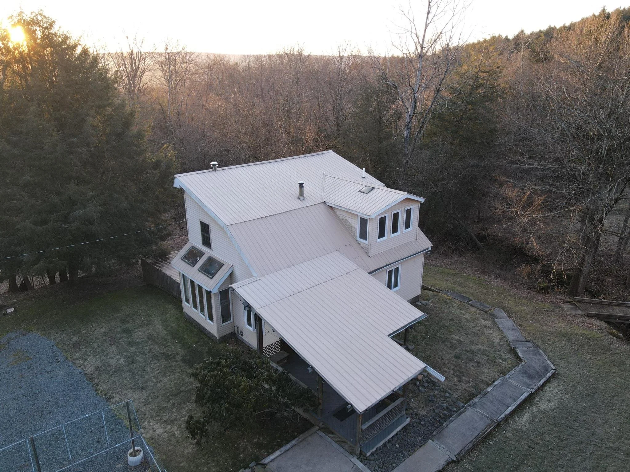 A house with a metal roof surrounded by trees and a fenced yard, seen from an aerial view during sunset.