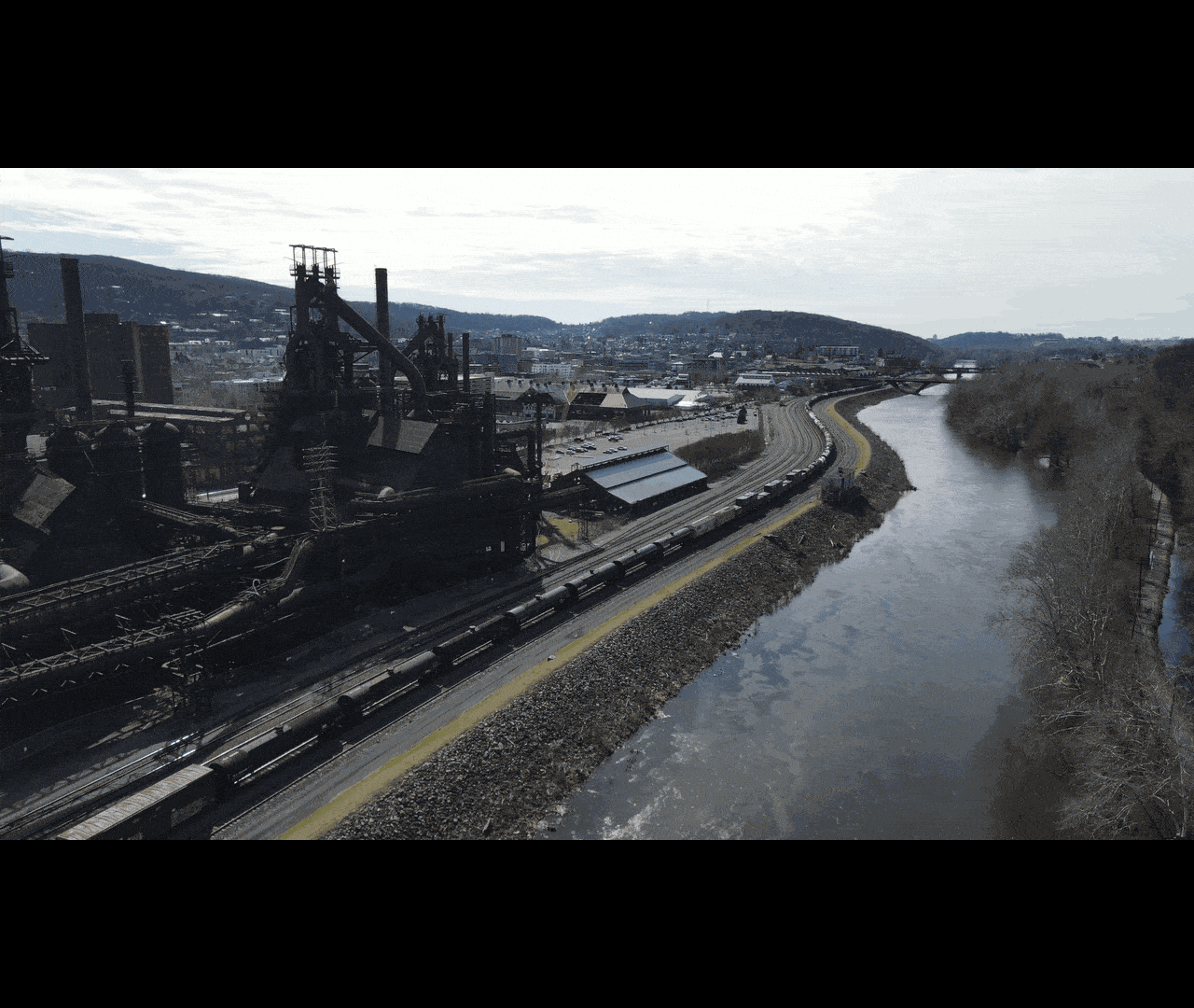 An industrial steel plant near a river with a highway running alongside in a cityscape with hills in the background.
