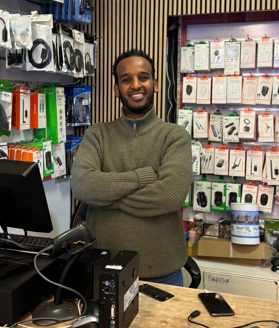 Man staat achter de toonbank van een elektronicawinkel, omringd door verschillende kabels en elektronische accessoires.