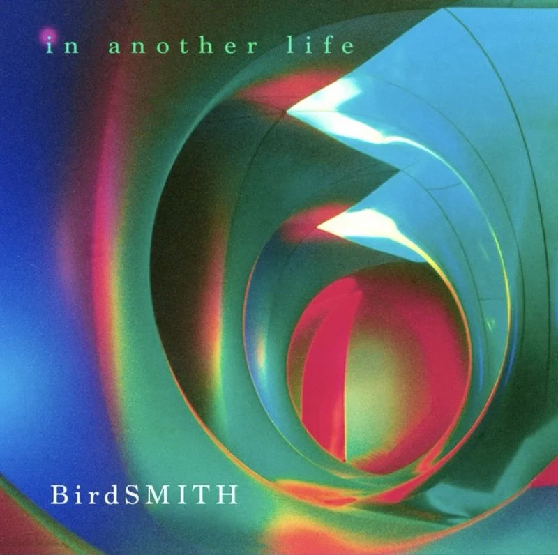 Abstract digital artwork with swirling green, red, and blue colors and the text 'in another life' at the top and 'BirdSMITH' at the bottom