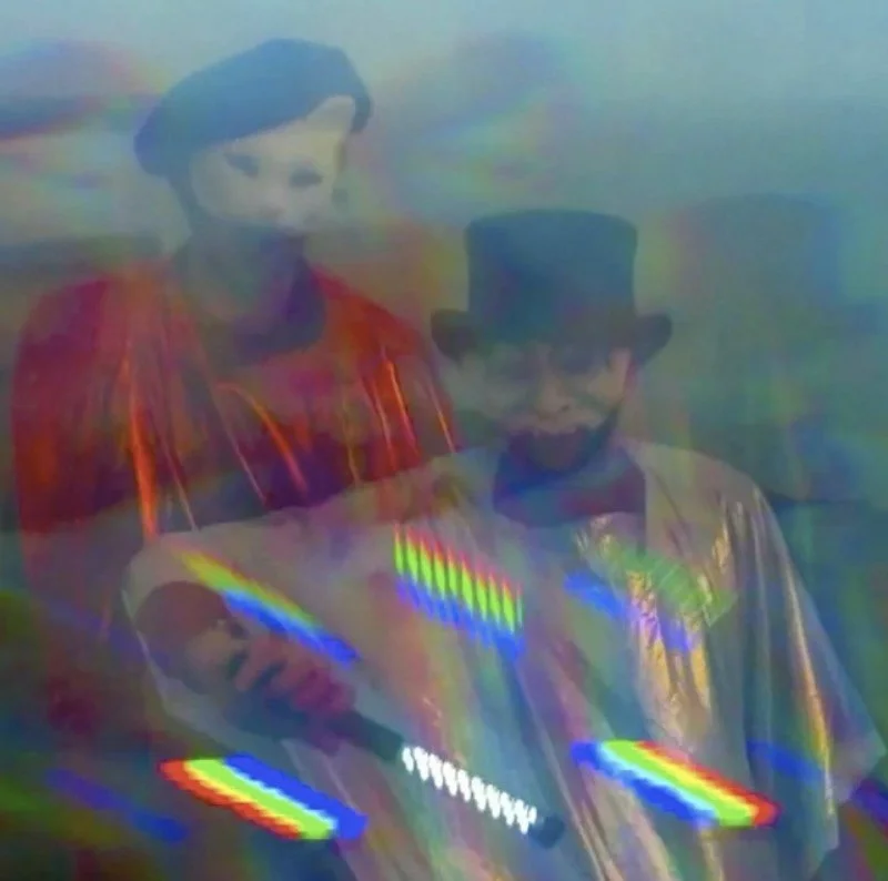 Two people with blurred, distorted appearance due to visual interference, one wearing a mask or mask-like face and a red shirt, the other wearing a top hat and sunglasses, dressed in a shiny or reflective jacket.