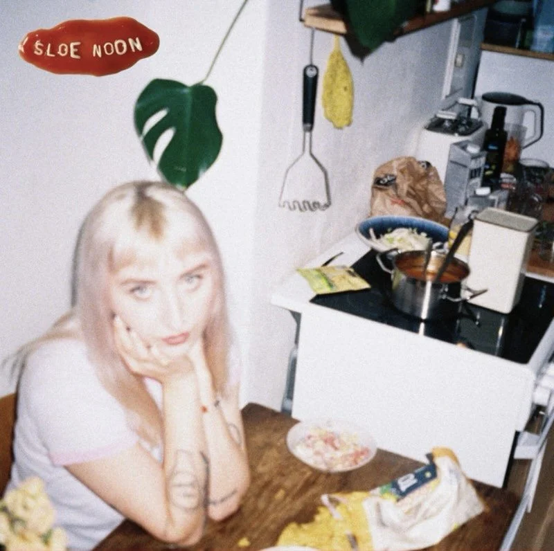 A woman with long blonde hair sitting at a table, resting her chin on her hands, in a kitchen with various cooking items and a green leafy plant on the wall.