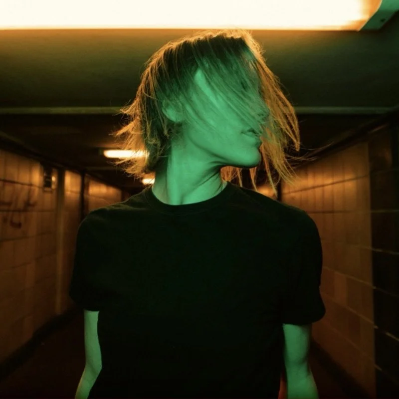 A person with shoulder-length blonde hair covering part of their face, wearing a black t-shirt, standing in a dimly lit tunnel with green and orange lighting.