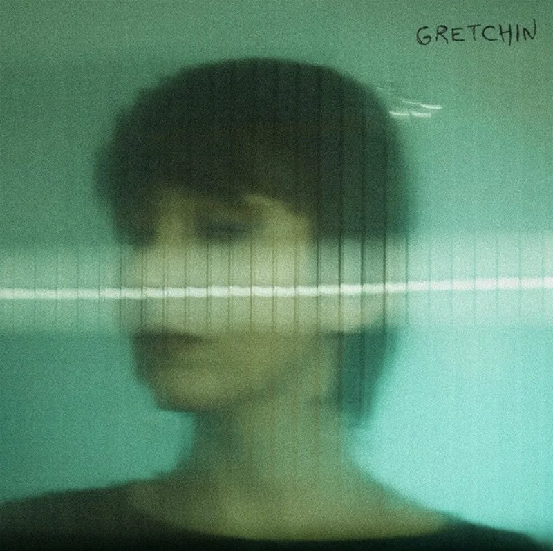 Blurred portrait of a woman with short dark hair wearing glasses, with a greenish background and the word 'GRETCHIN' written in the top right corner.