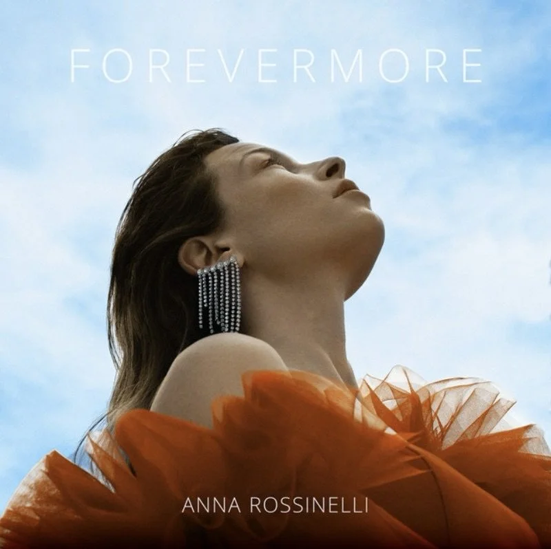 A woman with earrings and an orange dress, looking upward against a blue sky, with the text 'FOREVERMORE' and 'ANNA ROSSINELLI' overlaid.