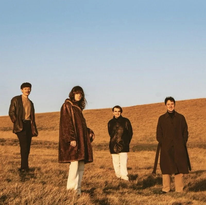 Four people standing on a grassy field during sunset or sunrise, with rolling hills and a clear blue sky in the background.