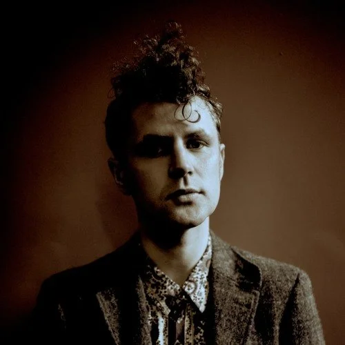 Portrait of a young man with curly hair, wearing a patterned shirt and a dark blazer, with a serious expression, standing against a dark background.