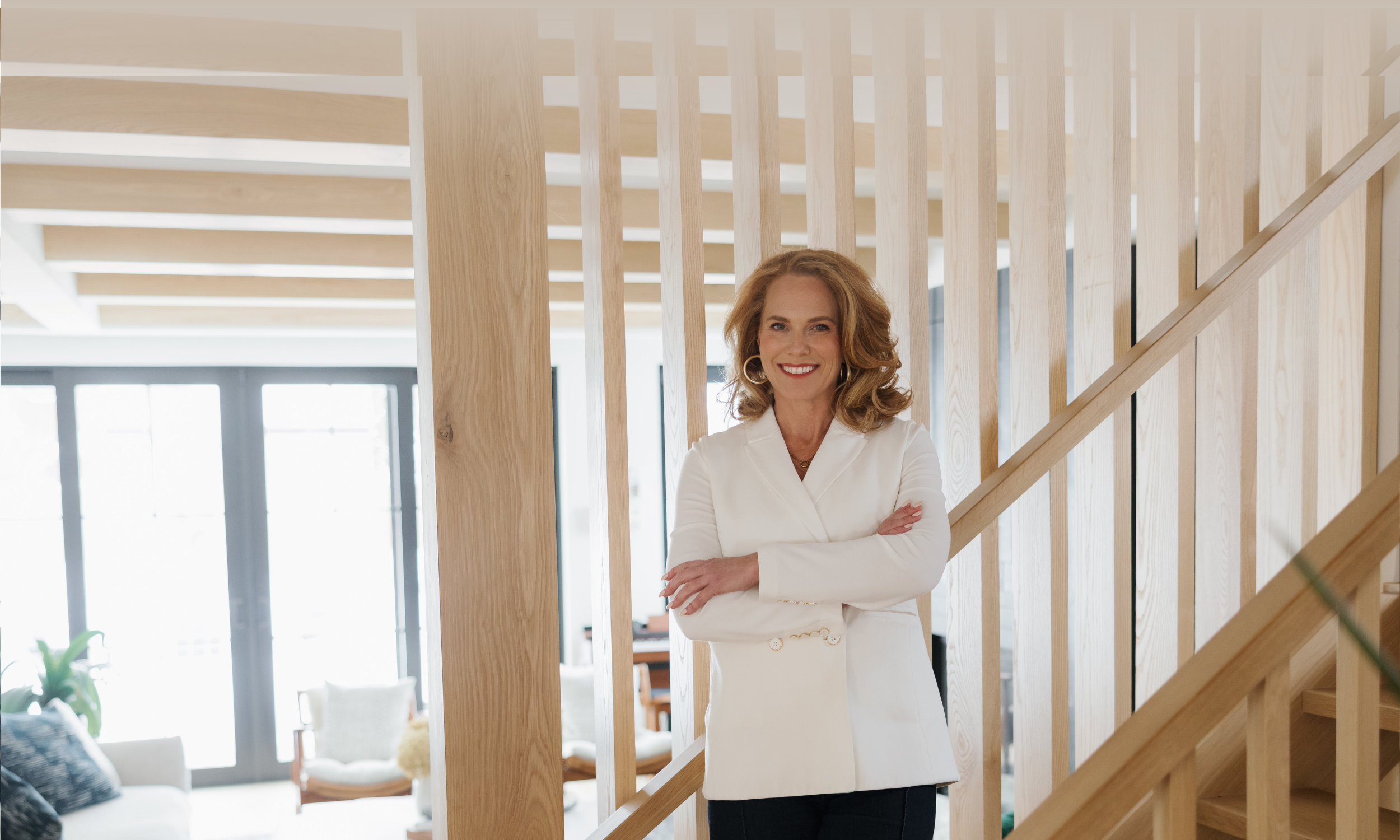 Woman with curly blonde hair wearing a white blazer smiling with arms crossed inside a modern home with wooden staircase and large windows in the background.