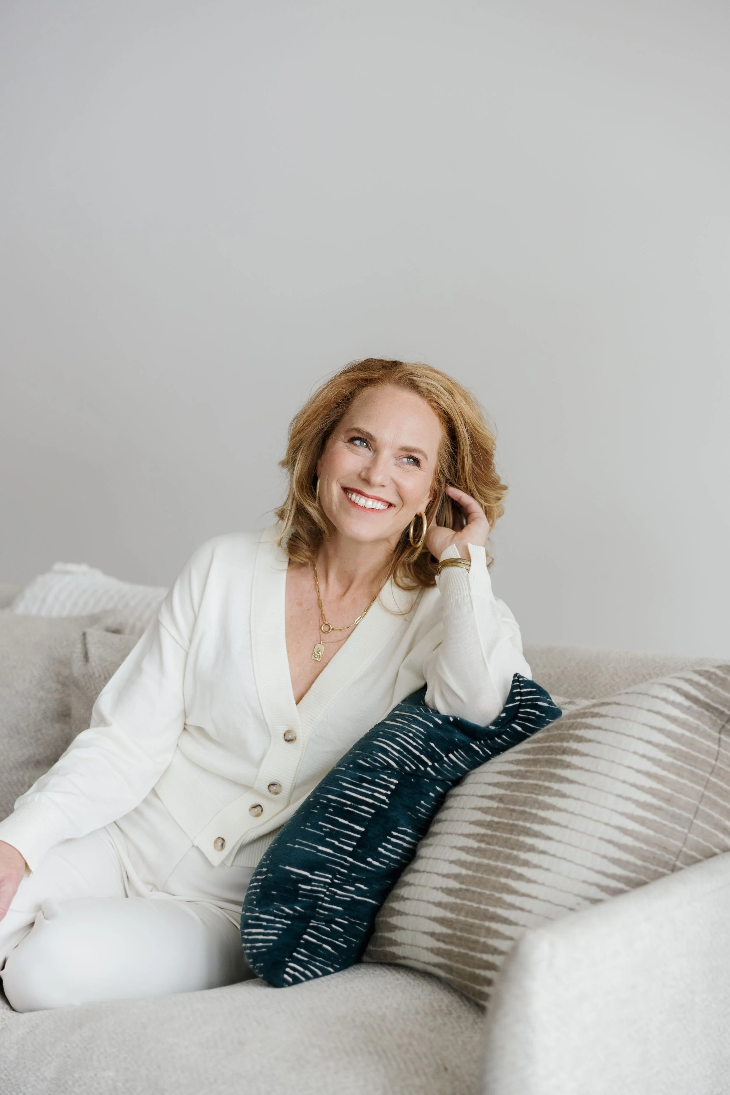 A woman with red hair, wearing a white cardigan and white pants, sitting on a beige sofa with patterned pillows, smiling and looking off to the side.