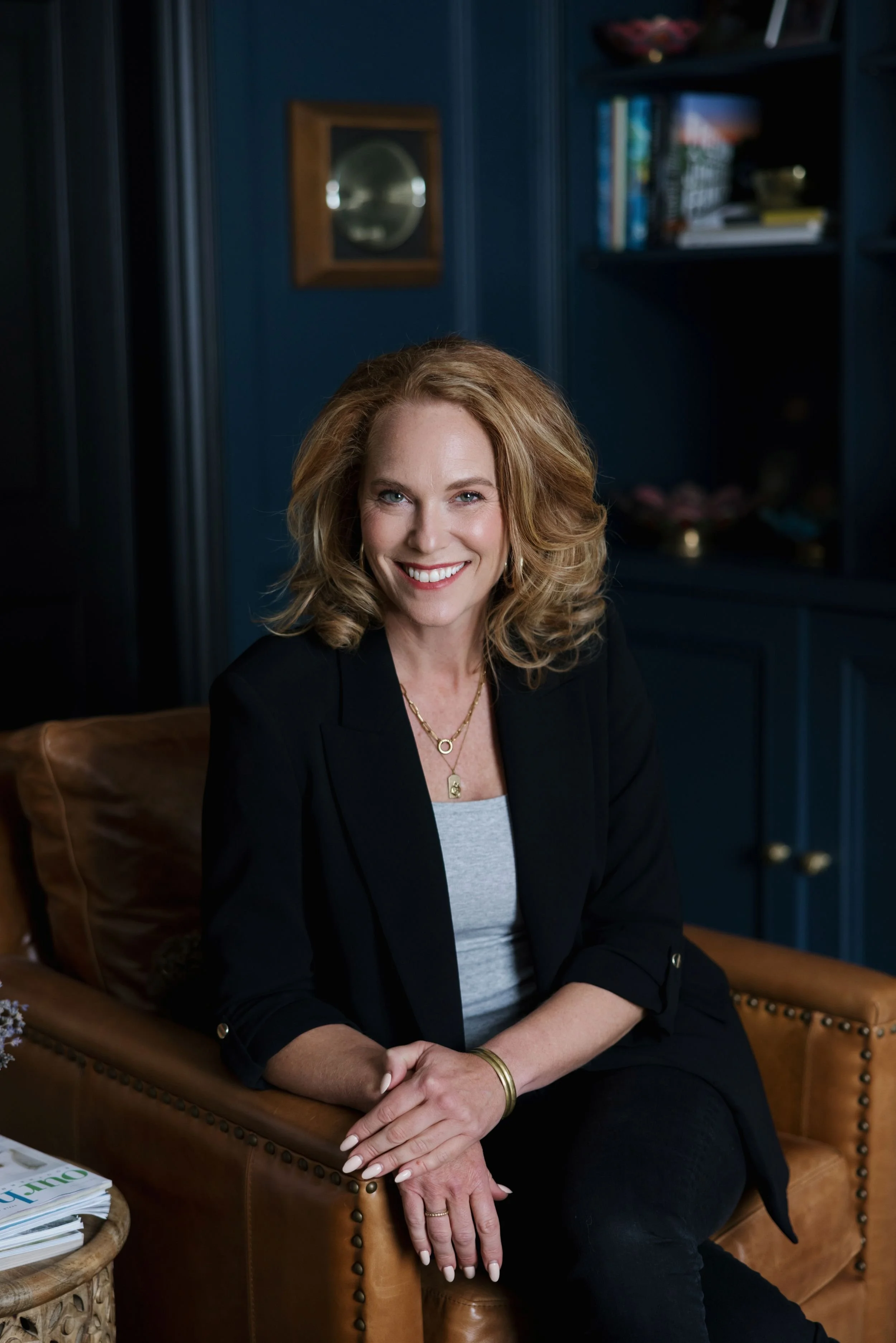 A woman with blonde, curly hair wearing a black blazer and gray top, sitting on a brown leather chair in a dimly lit room with navy blue walls, bookshelves, and decorative items.