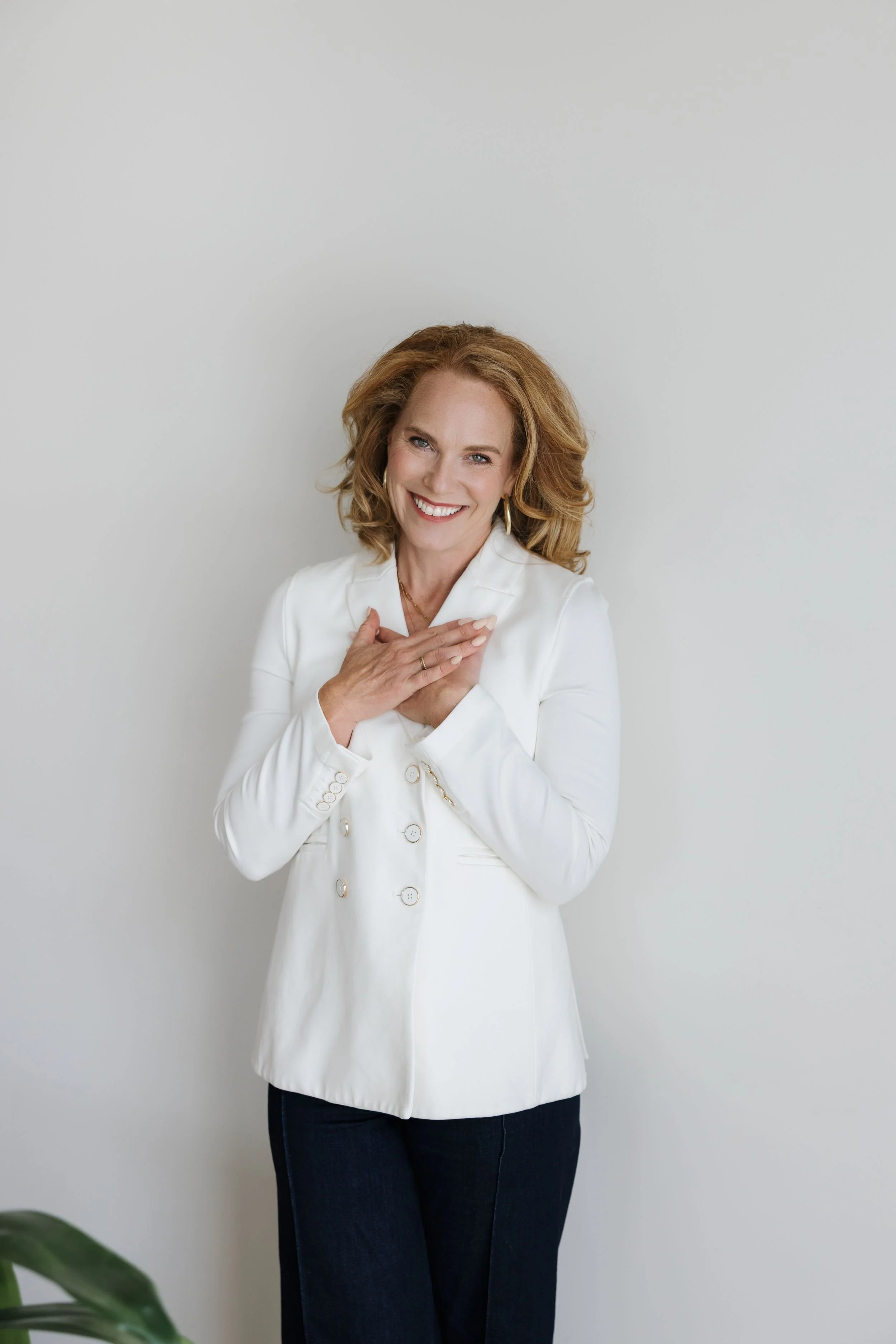Amy Halpenny, founder of Launchpad, with curly, shoulder-length hair smiling while wearing a white blazer and dark pants, standing against a plain white wall.