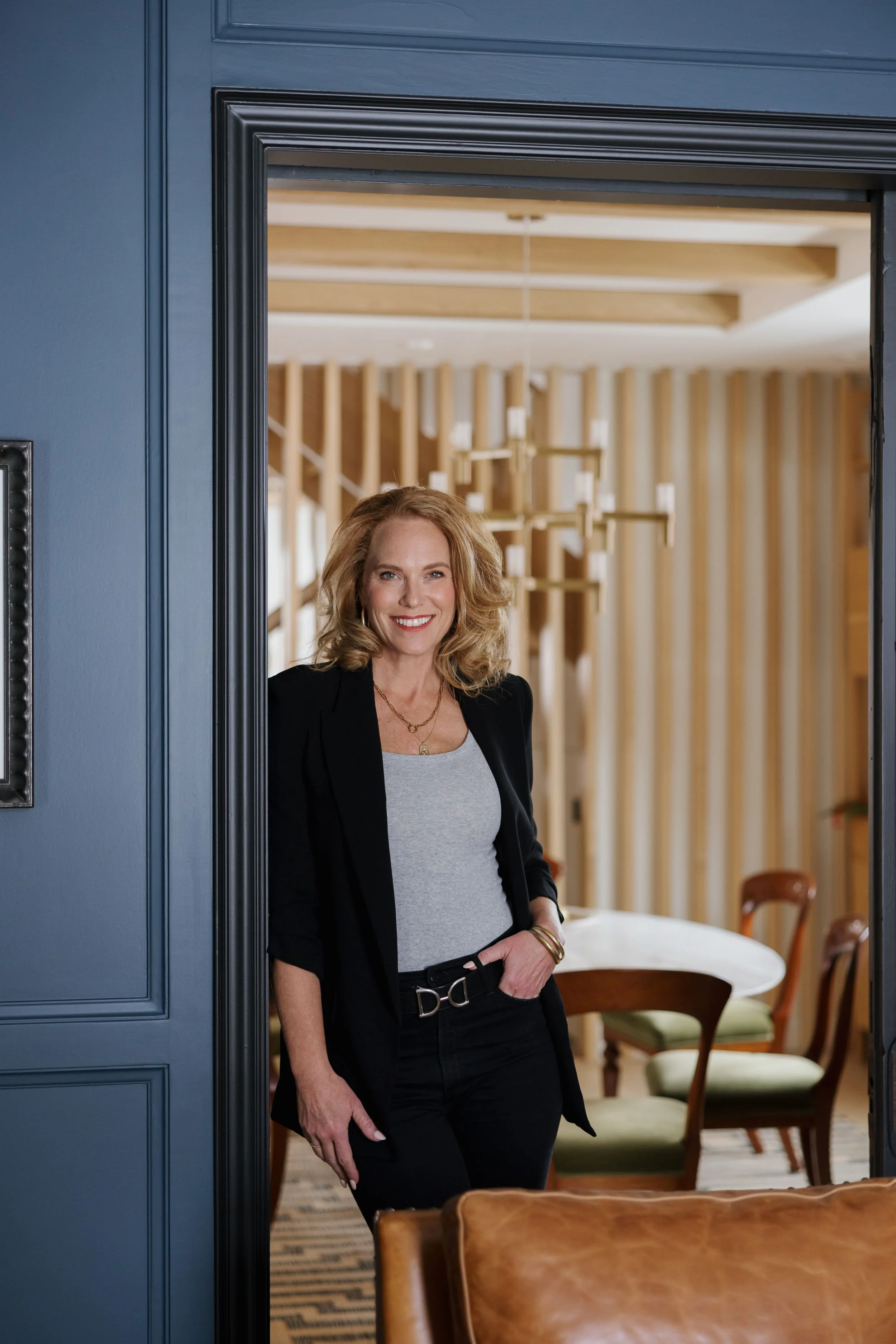 Portrait of Amy Halpenny of Launchpad Coaching + Advisory, standing in a doorway of a dining room with wooden chairs and a round table.