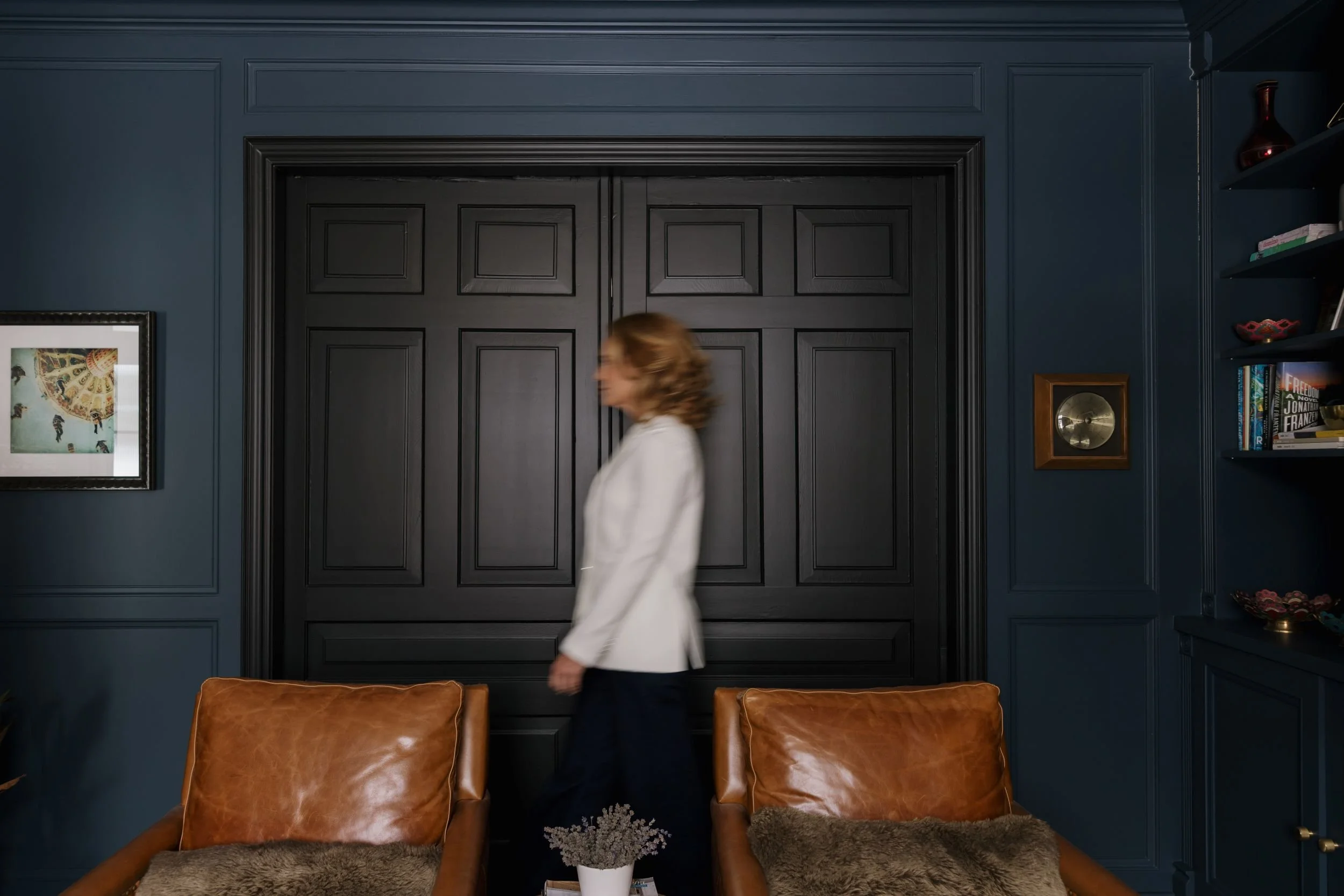 A woman in a white blazer and dark pants walking past black double doors in a blue-walled room, with leather chairs, a plant, a framed artwork, and a bookshelf visible in the room.