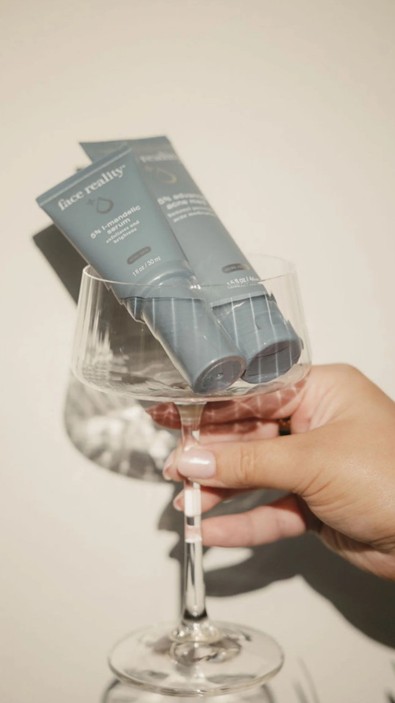 A hand holding a ribbed wine glass with two tubes of skincare products resting inside the glass.