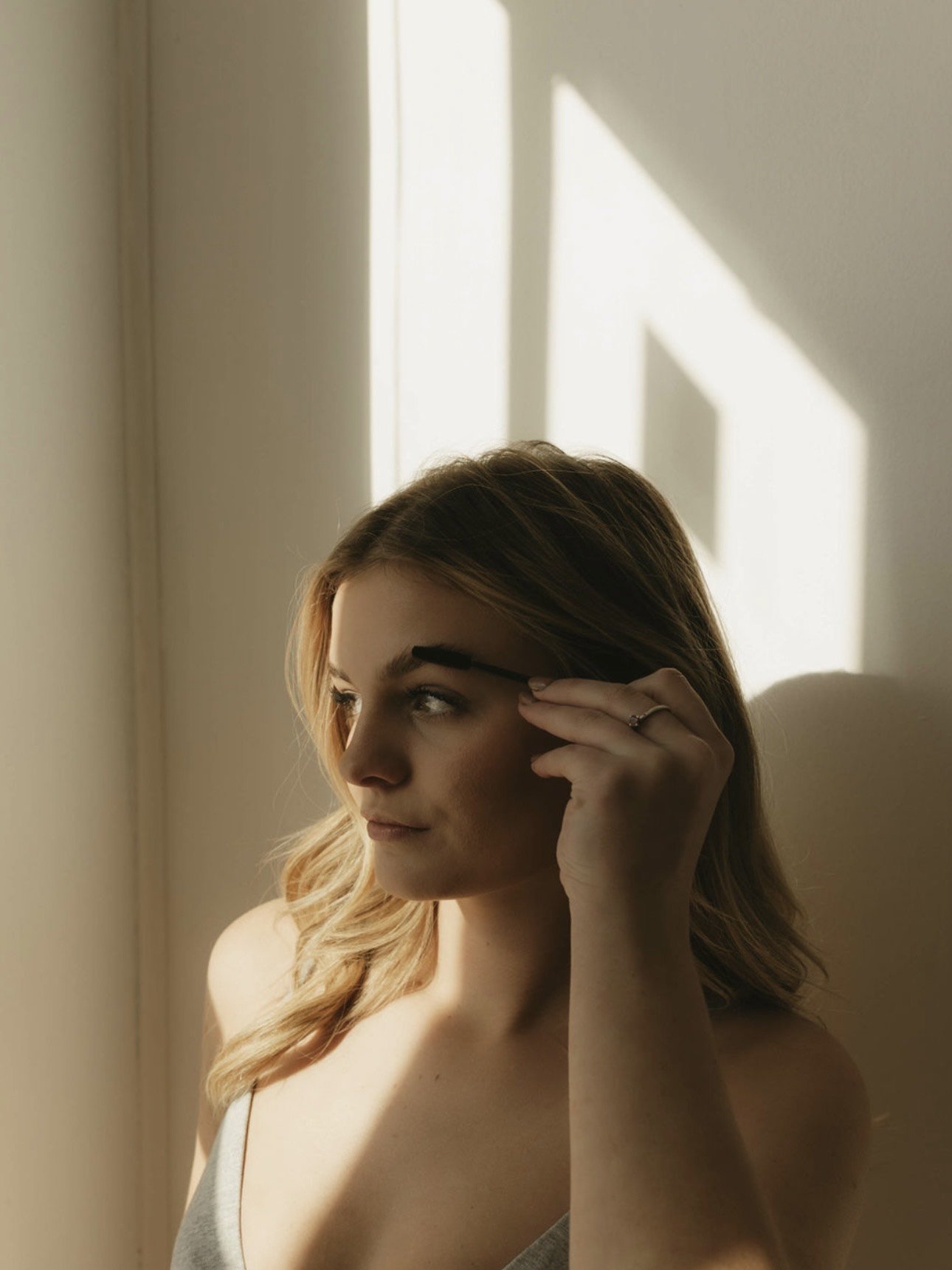 Woman with light brown hair, looking out a window with sunlight casting shadows on the wall, holding a eyebrow brush near her eyebrows.
