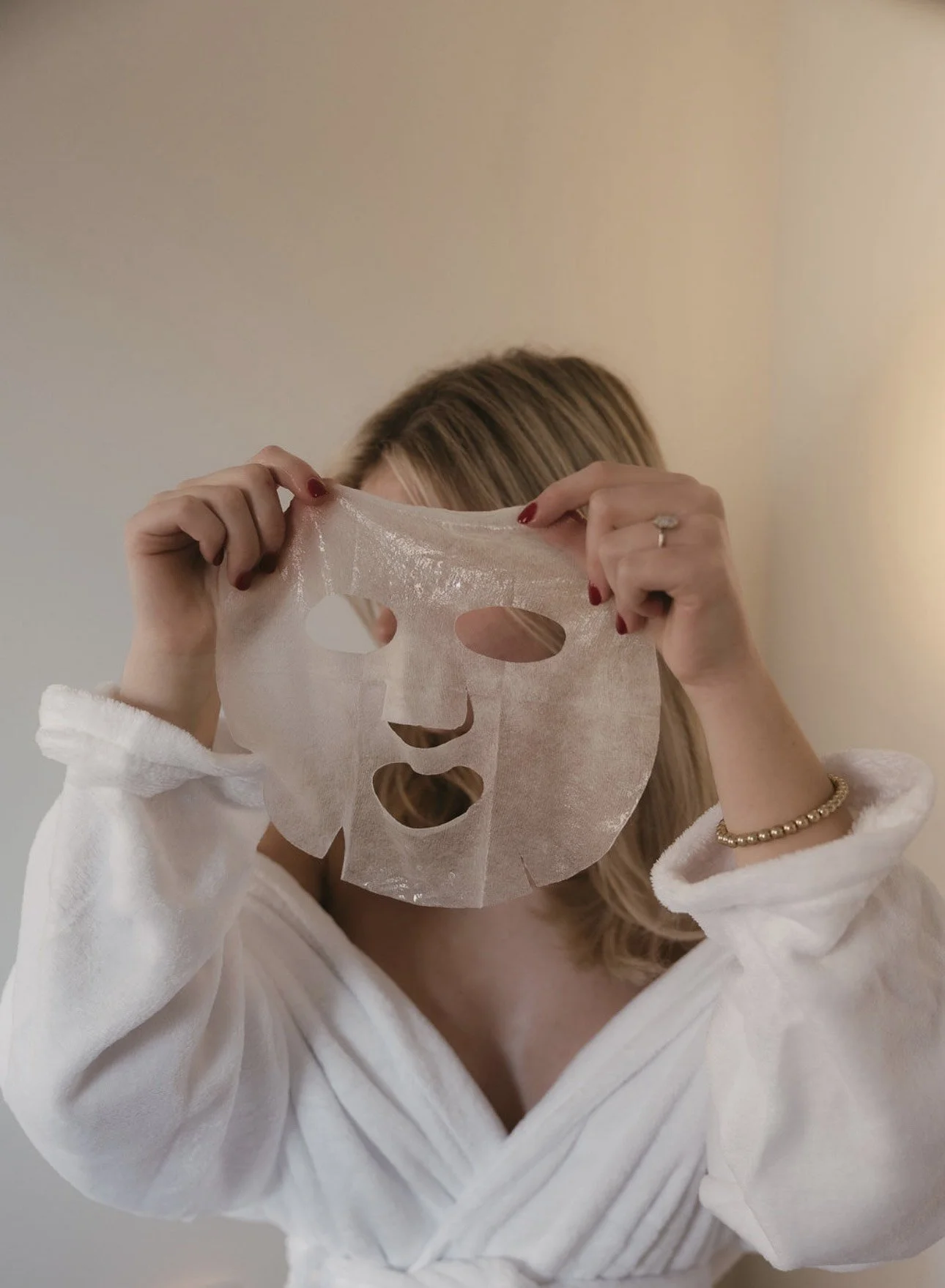 A woman wearing a white bathrobe is holding a sheet face mask in front of her face, covering it. The background is plain and neutral.