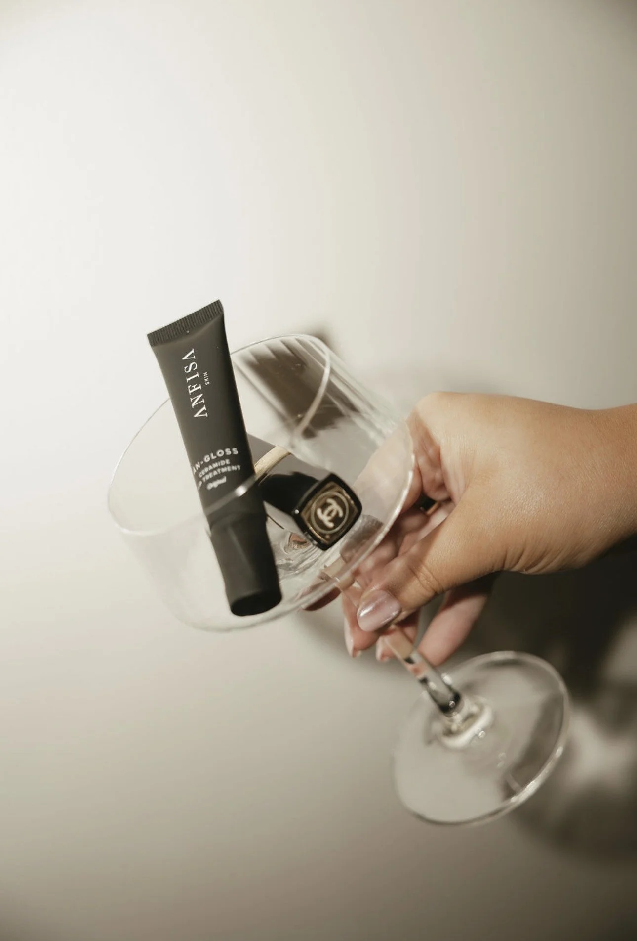 A person's hand holding a stemmed wine glass with a small black tube of lip treatment inside the glass.
