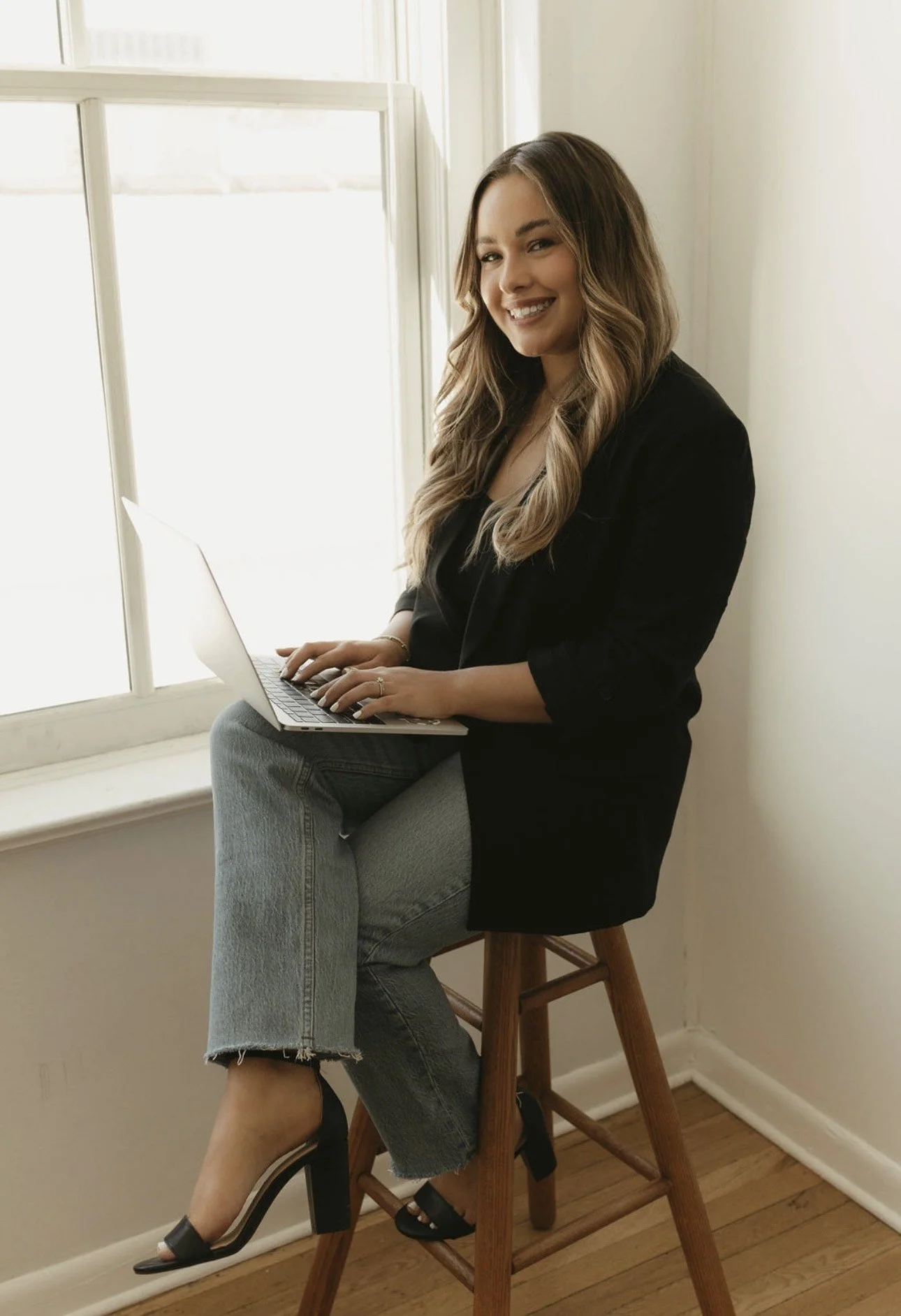 A woman sitting on a wooden stool near a window, using a laptop, smiling at the camera, wearing a black blazer, jeans, and black high heels.