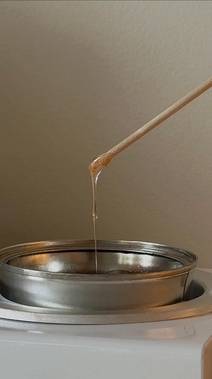 A close-up image of a wooden stick into a wax pot dripping wax into a metal container.