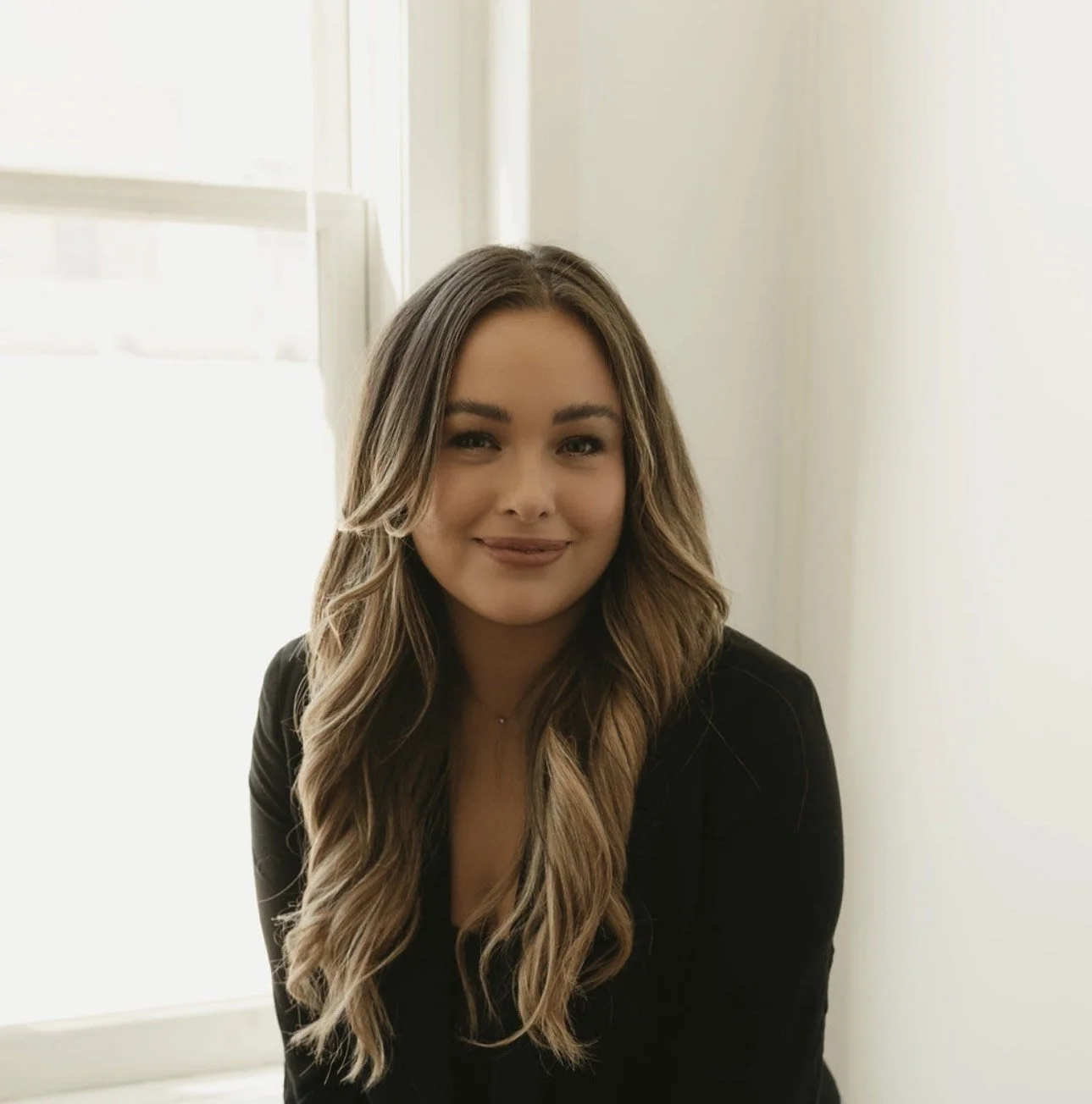 A young woman with long wavy hair smiling, sitting near a window with light coming through.