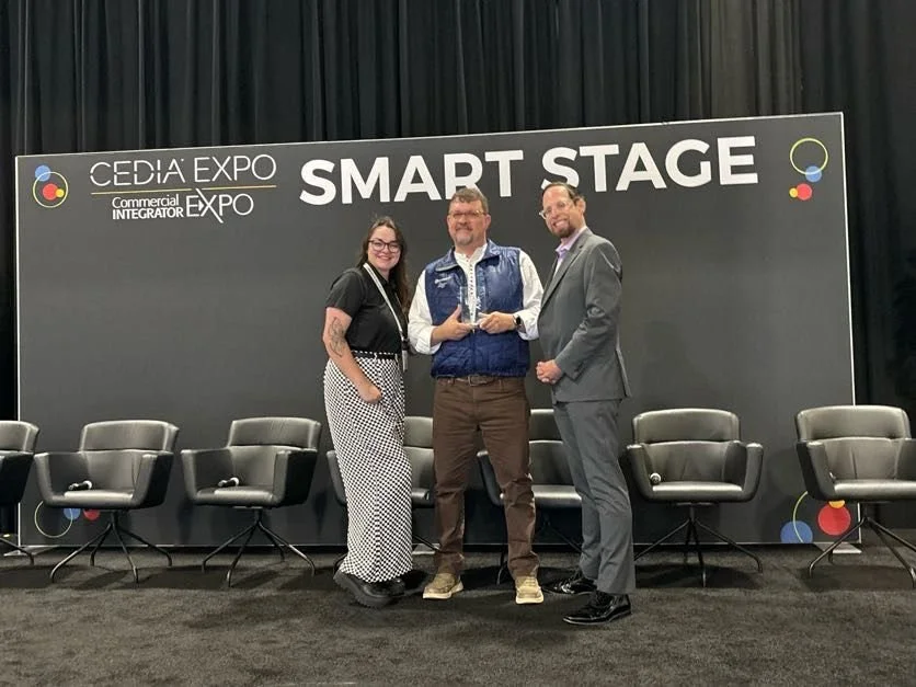 Karlie with two other people standing on a stage with a sign that reads 'CEDIA Expo', 'Commercial Integrator Expo', and 'Smart Stage'. They are smiling.