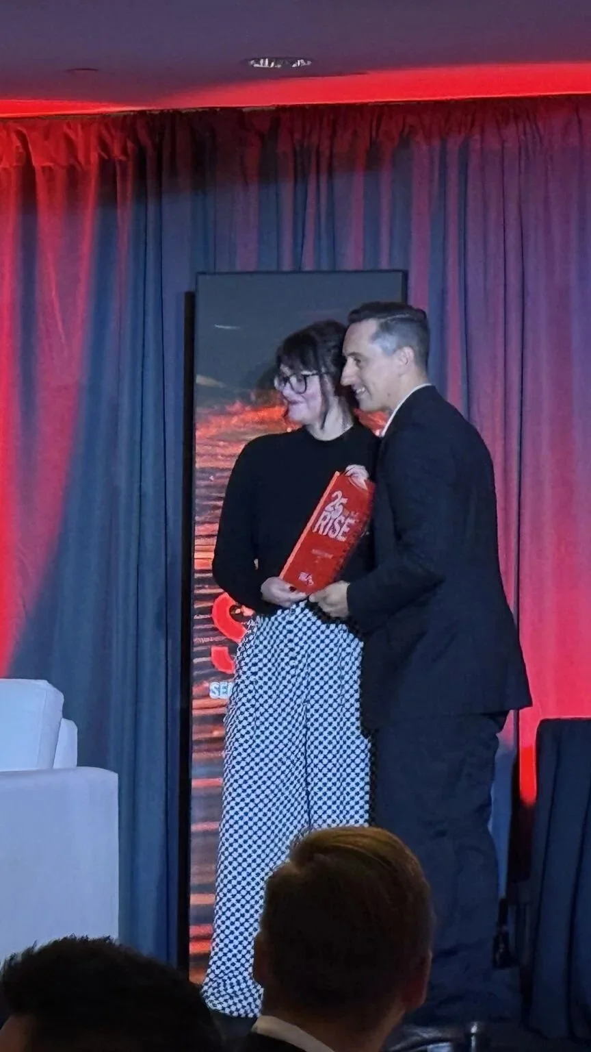 Karlie receiving the SIA 25 on the Rise award from a man on stage at an Securing New Ground, standing in front of red curtains, with a framed image of a sunset or sunrise behind them.