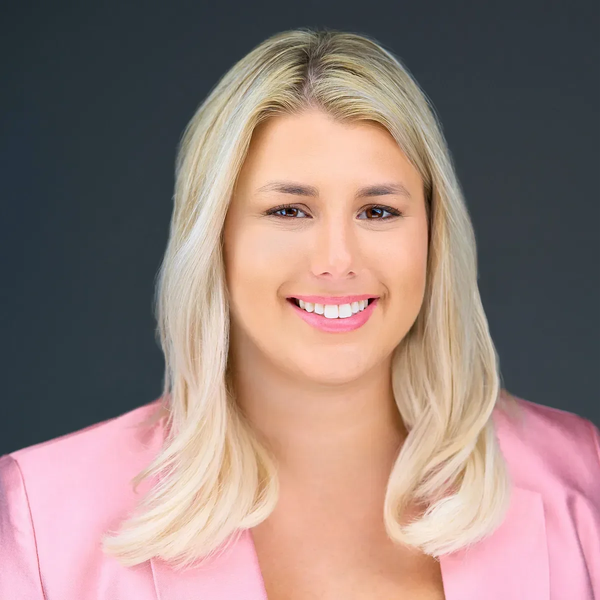 Headshot of a smiling woman with blonde hair wearing a pink blazer against a dark background.