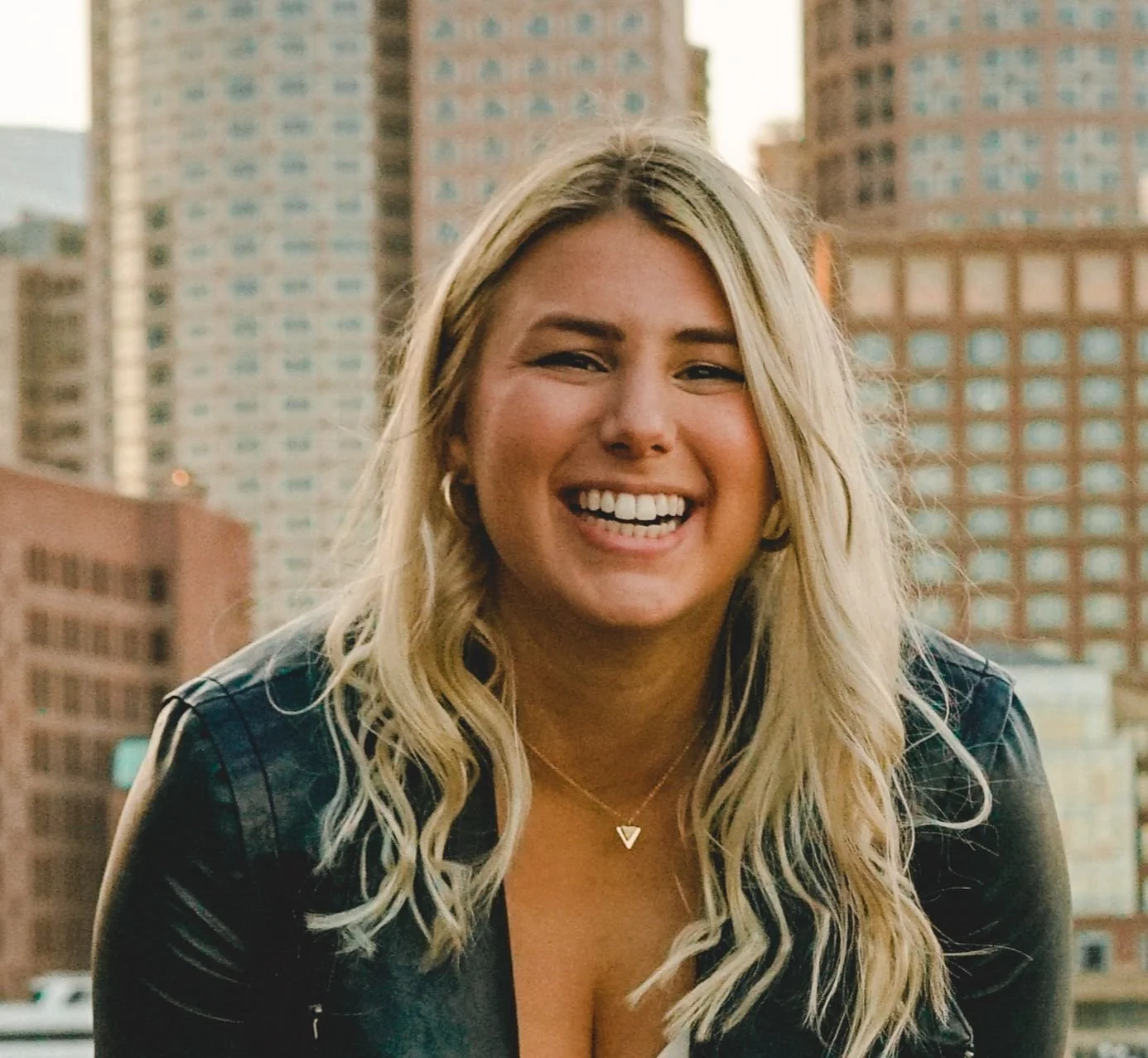 A smiling blonde woman with wavy hair, wearing a leather jacket, standing outdoors in front of tall city buildings.