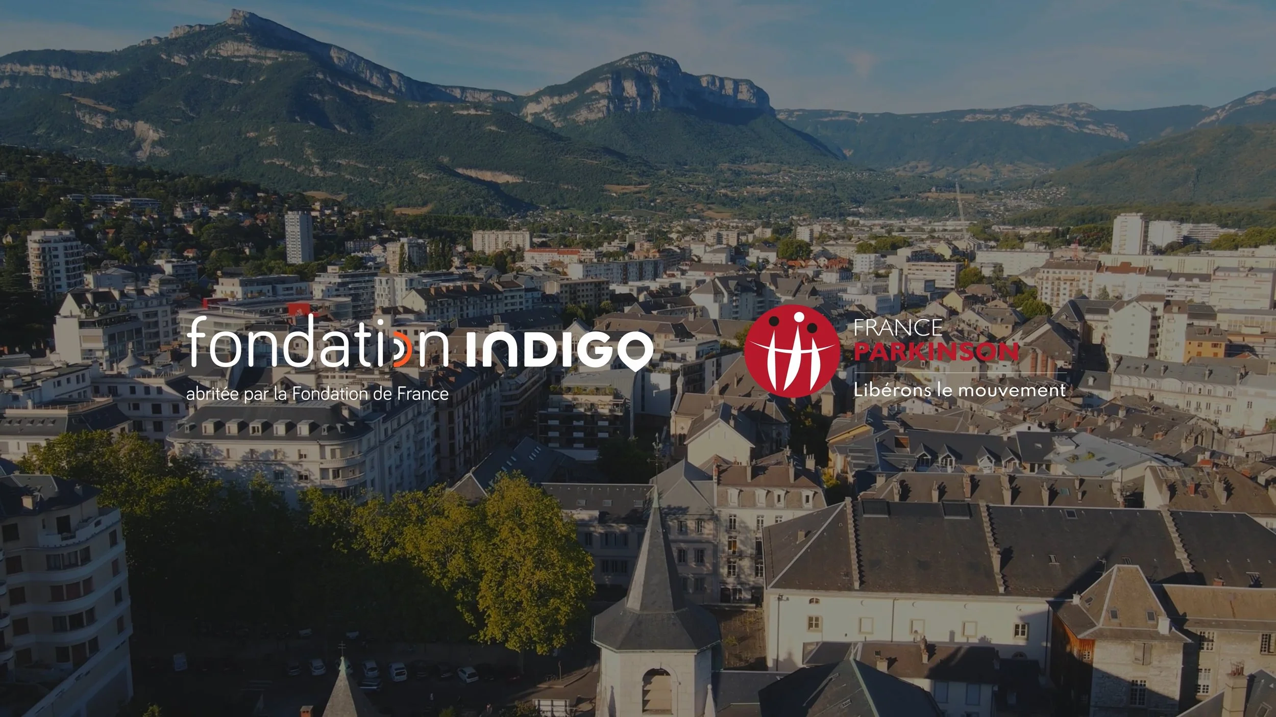 Vue aérienne d'une ville avec des bâtiments résidentiels et une montagne en arrière-plan, avec le logo de Fondation Indigo et de France Parkinson.