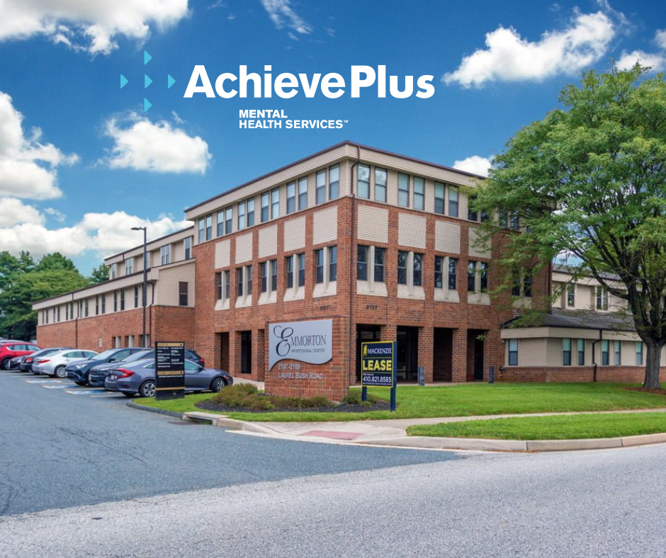 A multi-story brick building under a blue sky with white clouds, with parking lots and green trees surrounding it. Signs indicate the building houses a mental health services center called AchievePlus, with offices for Emorton and lease information for Mackenzie.