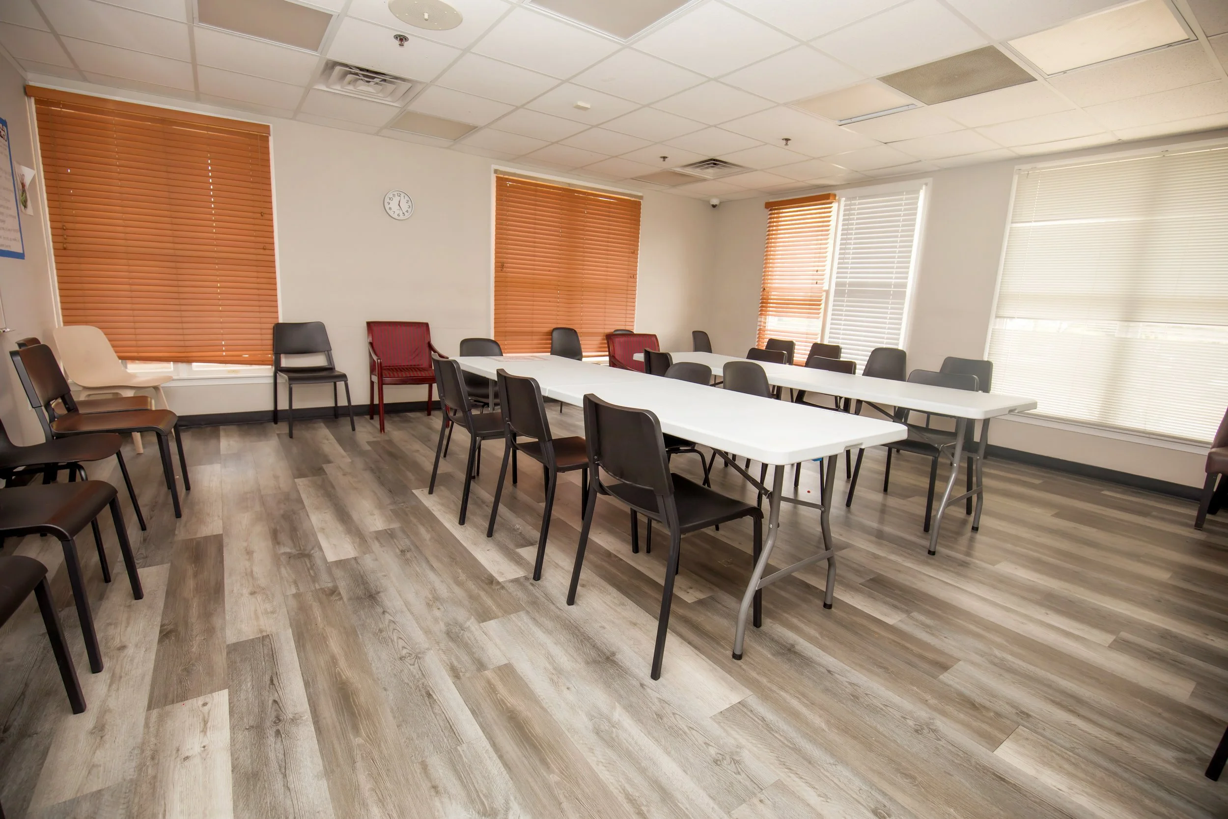 Empty conference room with long white tables, black and white chairs, orange and white blinds on windows, and a wall clock.