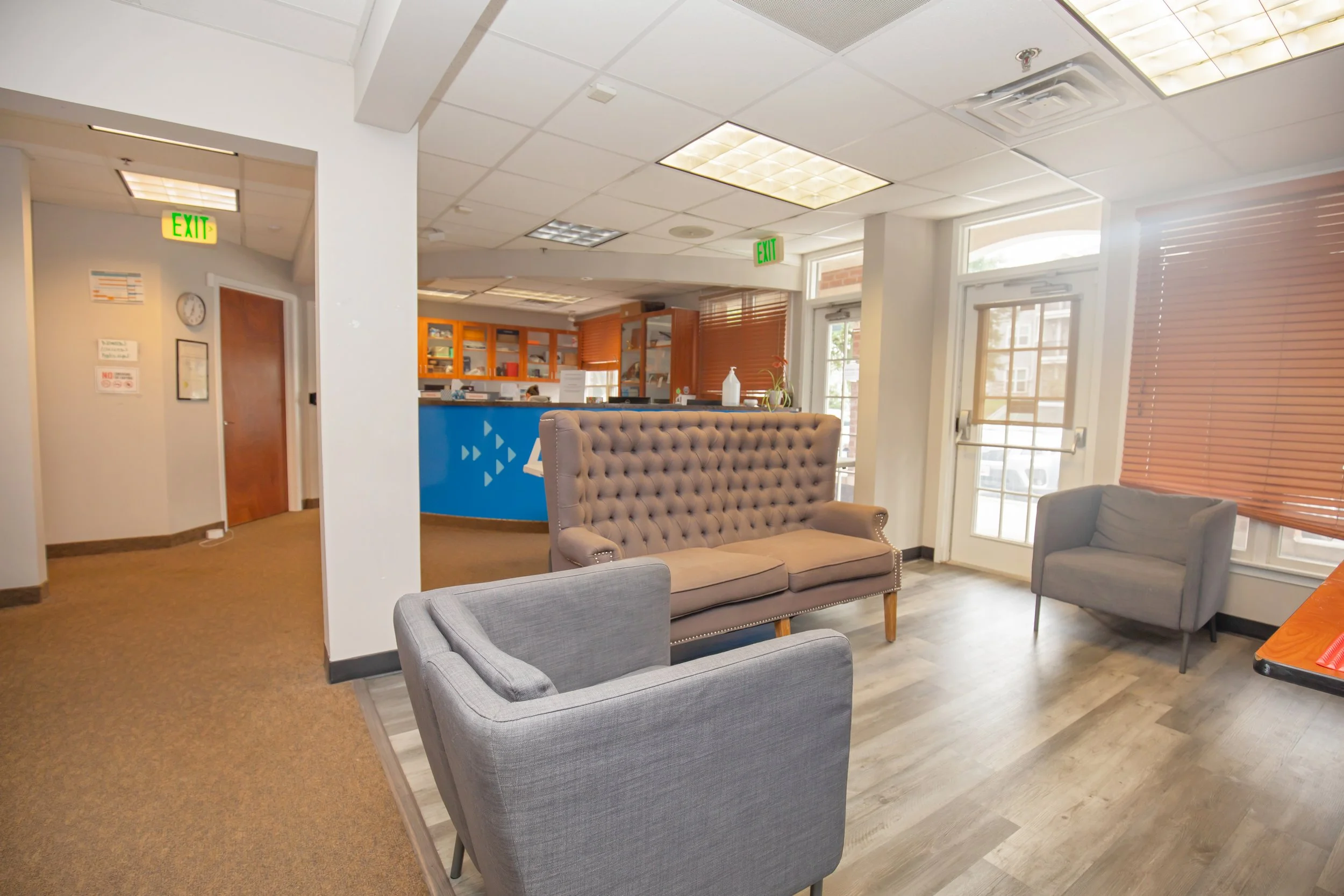 Waiting area with three chairs, a tufted sofa, a doorway with glass doors, and large windows with blinds in a healthcare or office building.