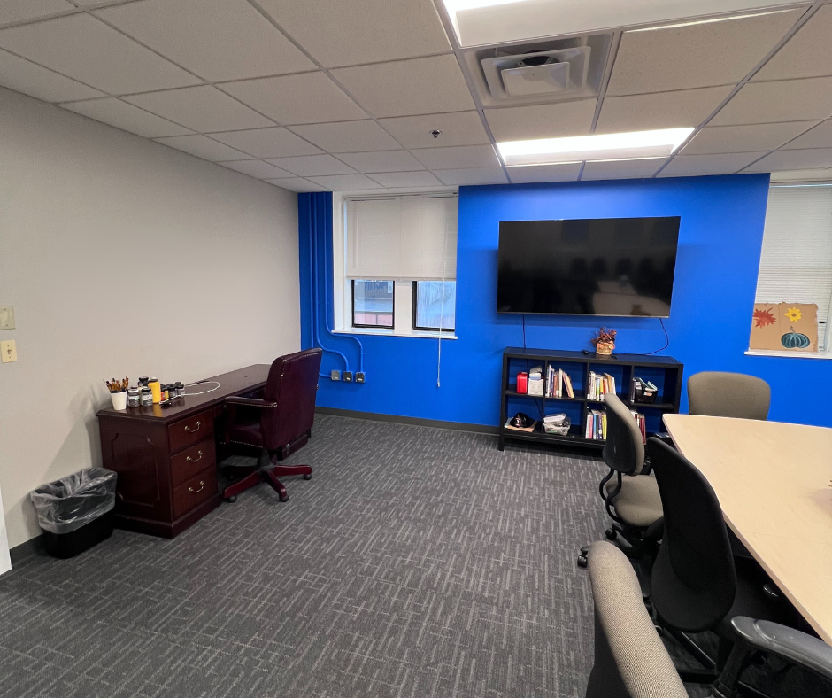 Empty conference room with long white tables, black and white chairs, orange and white blinds on windows, and a wall clock.