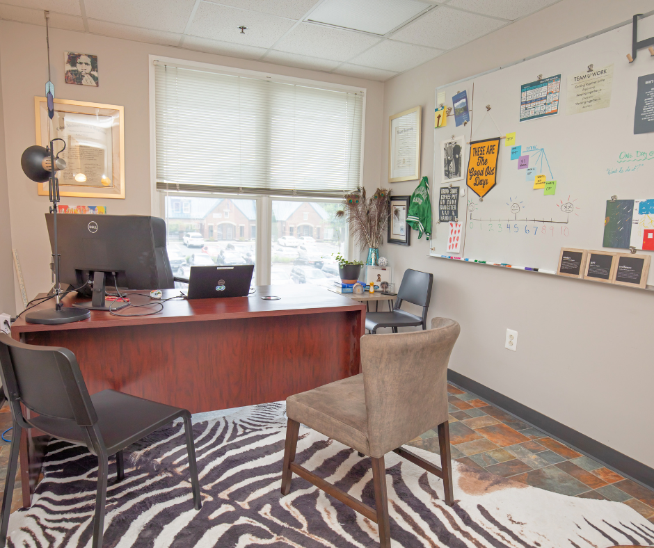 An office with a large wooden desk, three chairs, a whiteboard with notes, and a window with blinds overlooking parked cars and houses.