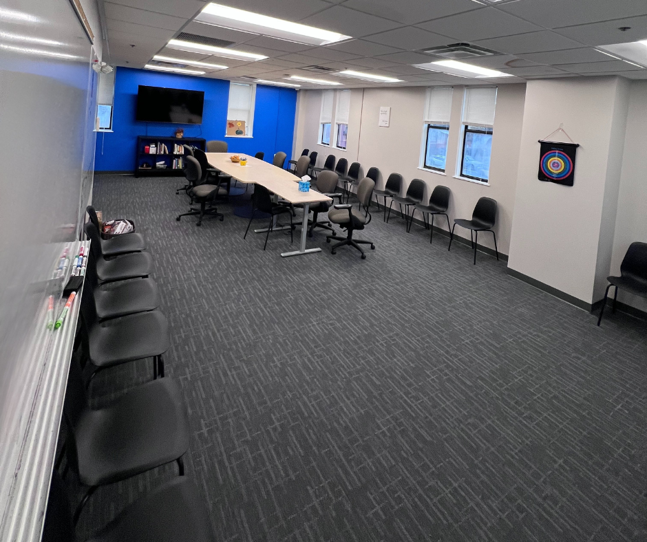Empty conference room with rows of black chairs, a long table, a large screen TV, windows, and a whiteboard.