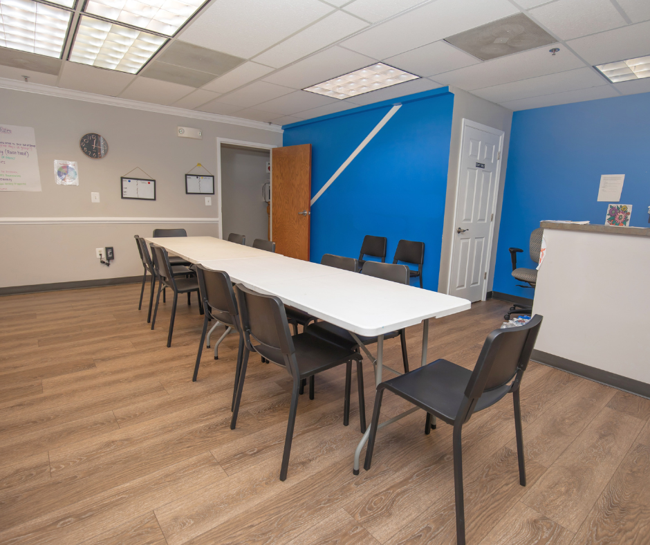 Meeting room with a long white table, multiple black chairs, a blue accent wall, a closed white door, and framed items on the wall.
