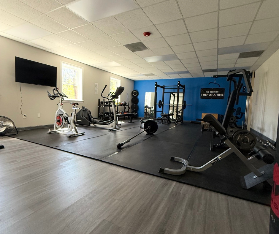 Empty gym with workout equipment including stationary bike, weightlifting bench, free weights, and a cable machine, with a television on the wall and windows letting in natural light.