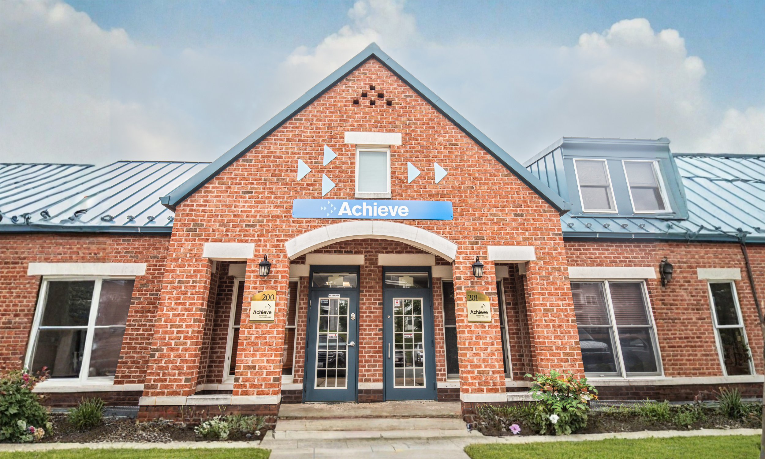 A brick building with two entrances, each having a sign with the word 'Achieve' and an address number. The building features a gabled roof with a small window near the top, and decorative elements including arrows and the word 'Achieve' above the entrance. There are well-maintained bushes and flowers in front of the building under a partly cloudy sky.