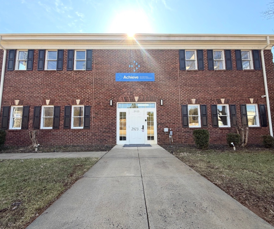 Front view of Achieve NC with a white door, black shutters on windows, and a blue sign that reads 'Achieve' with the logo of a healthcare organization, under clear skies with sunlight shining.