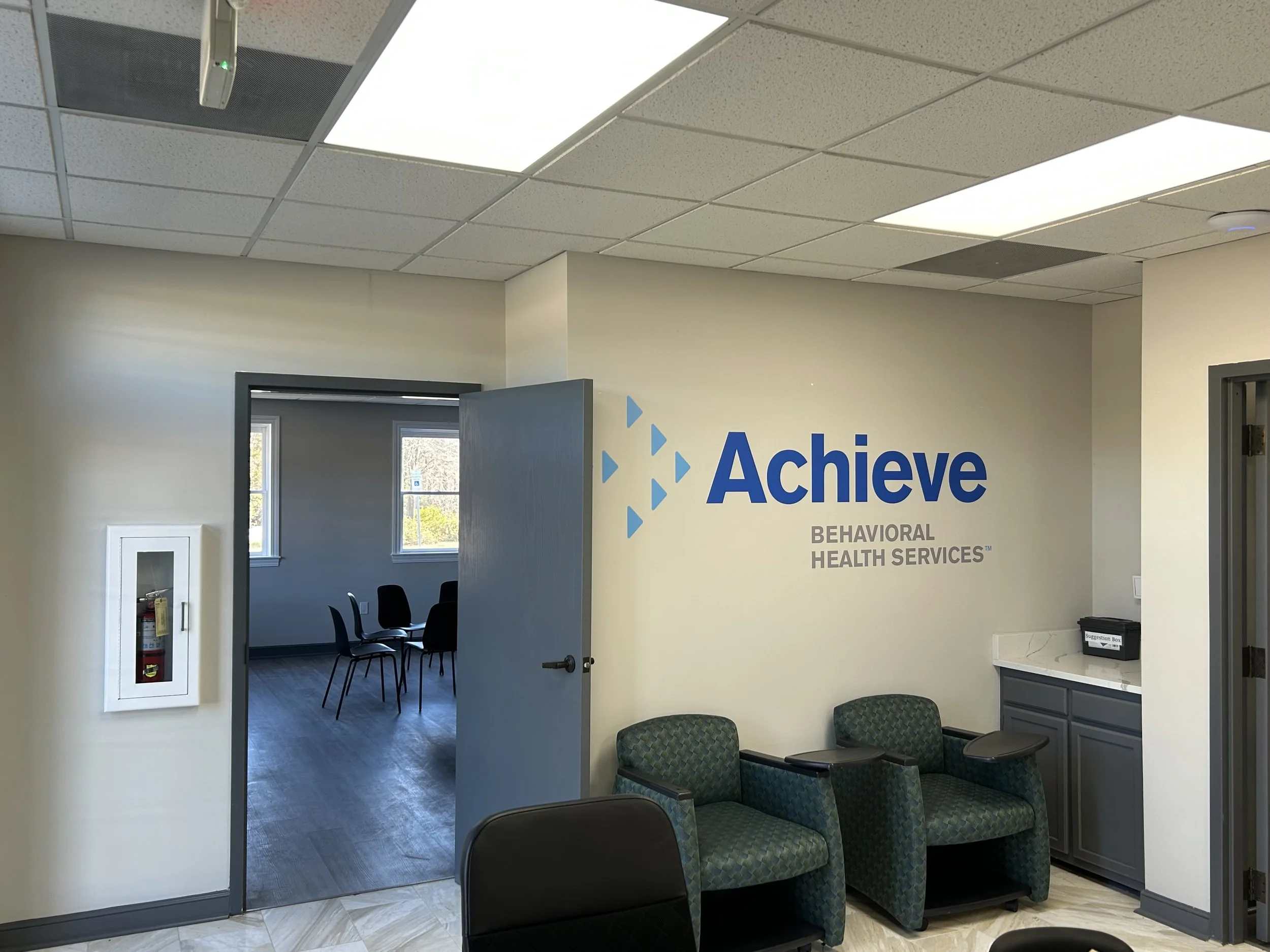 Interior of NC Achieve behavioral health services office with a wall sign reading 'Achieve Behavioral Health Services,' green patterned armchairs, a small counter with a suggestion box, and a doorway leading to a room with chairs and windows.
