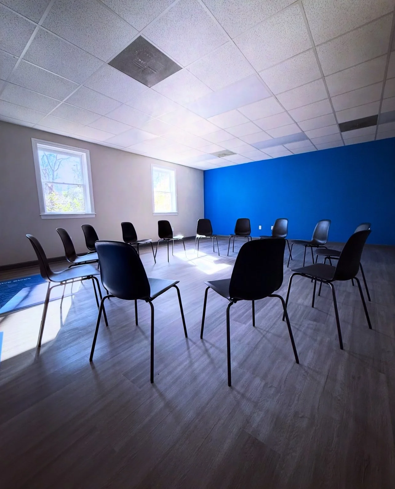 A room with a blue accent wall and two windows, featuring chairs arranged in a circle, likely set up for a meeting or group discussion.