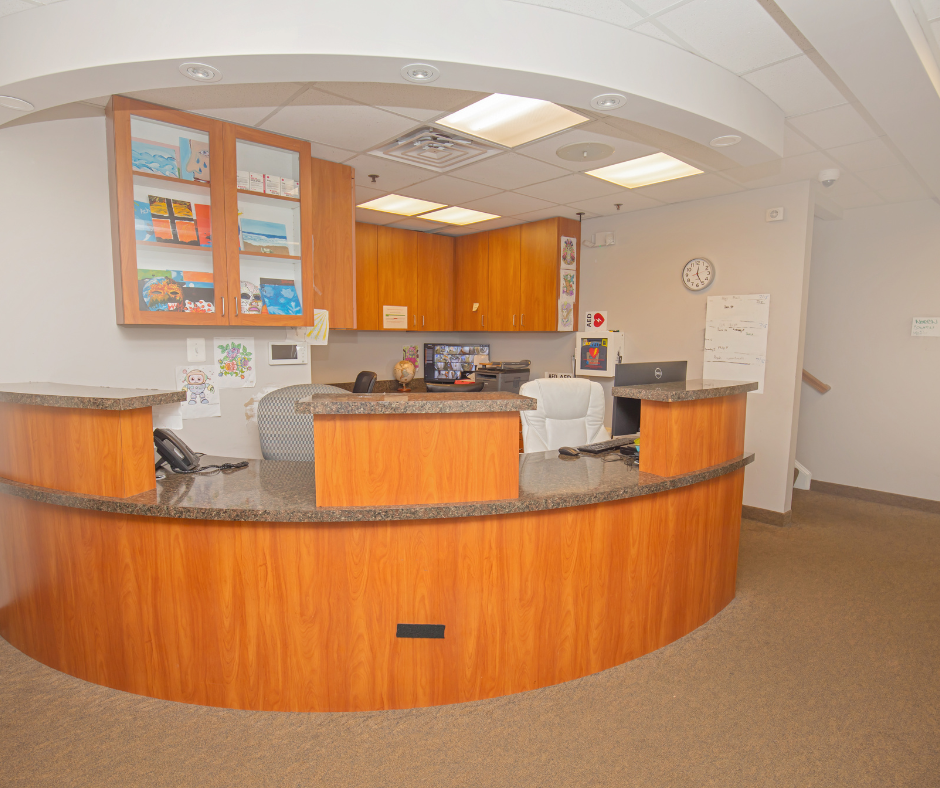 Reception desk area with wooden counter, white office chair, computer, and wall-mounted cabinets in a healthcare or office setting.