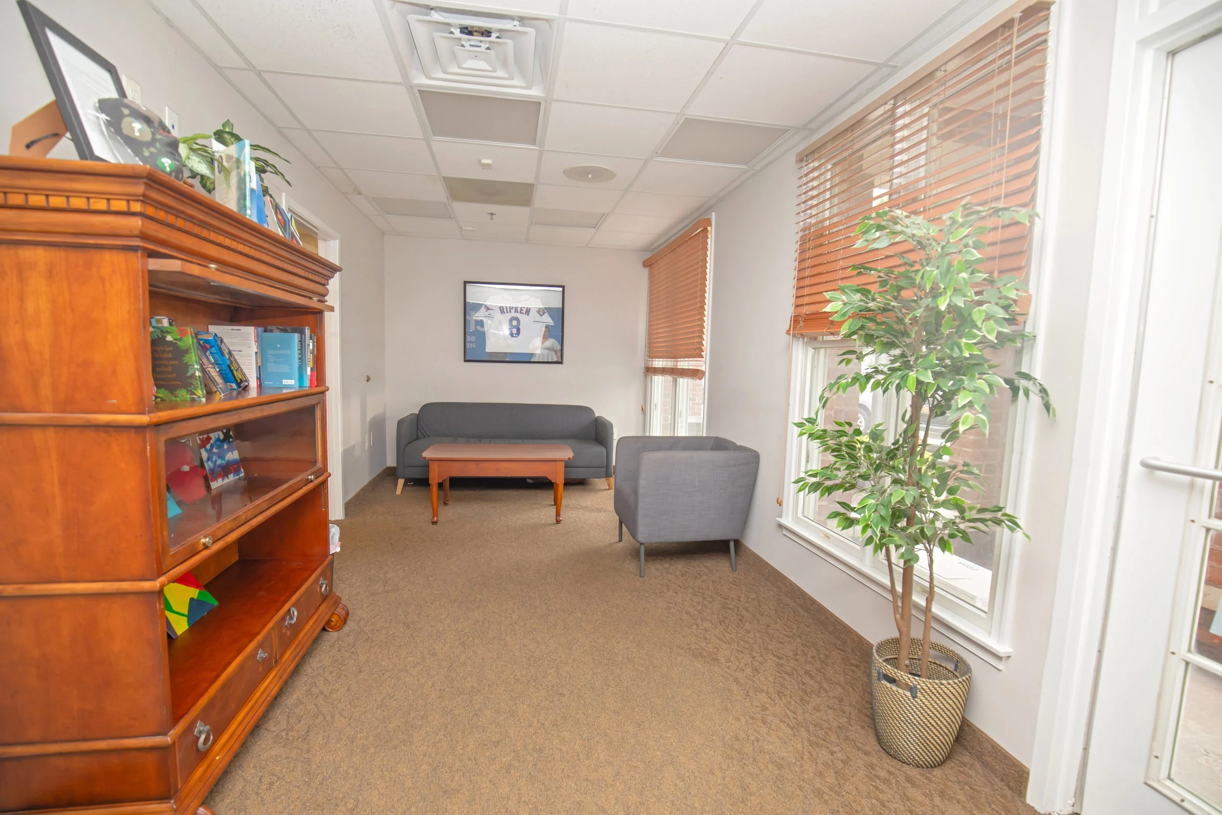 Empty office room with gray sofa, gray armchair, wooden coffee table, wooden bookshelf filled with books and decorative items, large window with brown blinds, potted plant, framed sports jersey on the wall, and white walls.