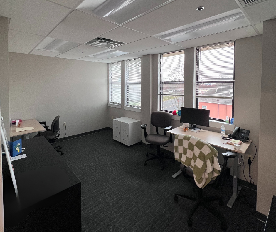 Group room with a table covered with an art project, chairs around, a large colorful wall hanging with the word 'achieve,' a whiteboard, a window with blinds, and a small cabinet with a computer on top.