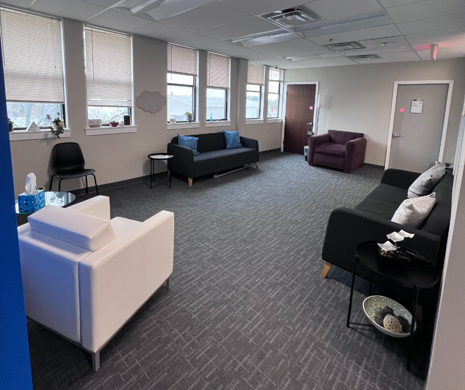A spacious waiting room with multiple sofas and chairs, large windows with blinds, and various decorative items.