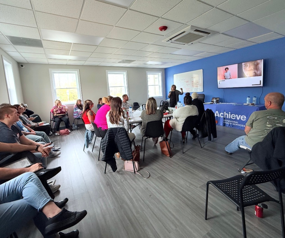 A woman giving a presentation to a group of people seated in a conference room with blue accent wall and a large screen.