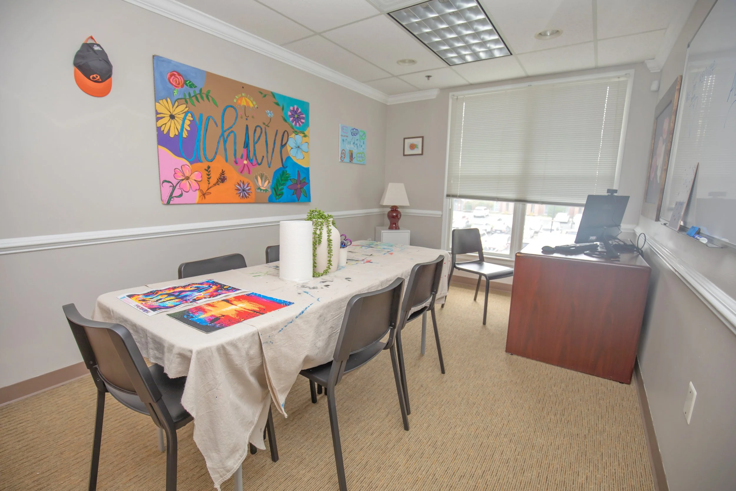 Group room with a table covered with an art project, chairs around, a large colorful wall hanging with the word 'achieve,' a whiteboard, a window with blinds, and a small cabinet with a computer on top.