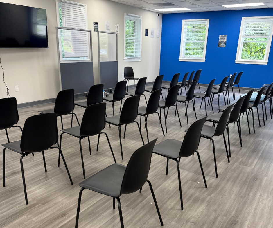 Room with multiple rows of black chairs facing a large flat-screen TV on the left wall. The room has white and blue walls, three windows with white blinds, and a wooden floor.