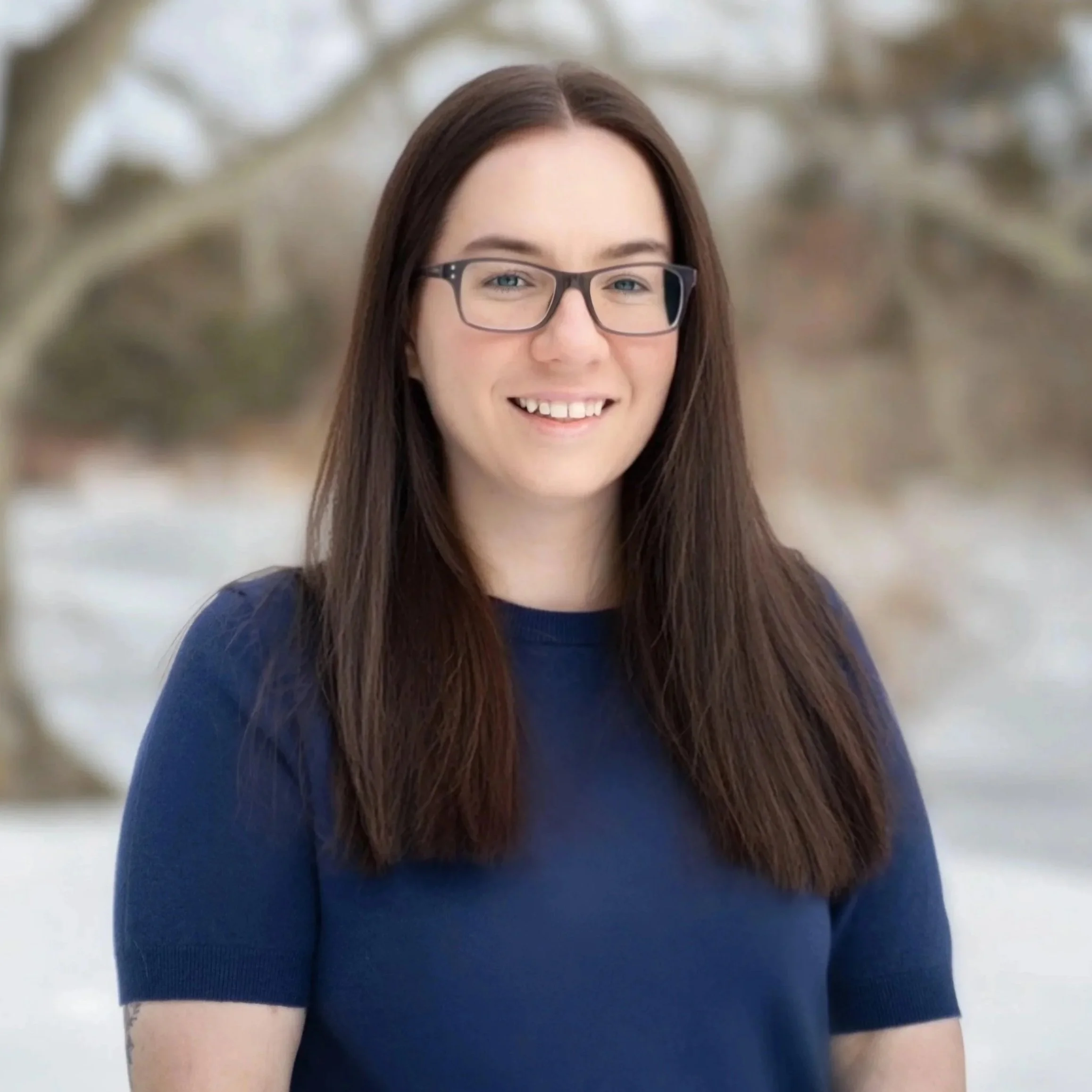Portrait of a woman with blonde hair, wearing a white shirt and hoop earrings, smiling against a blurred beige background.