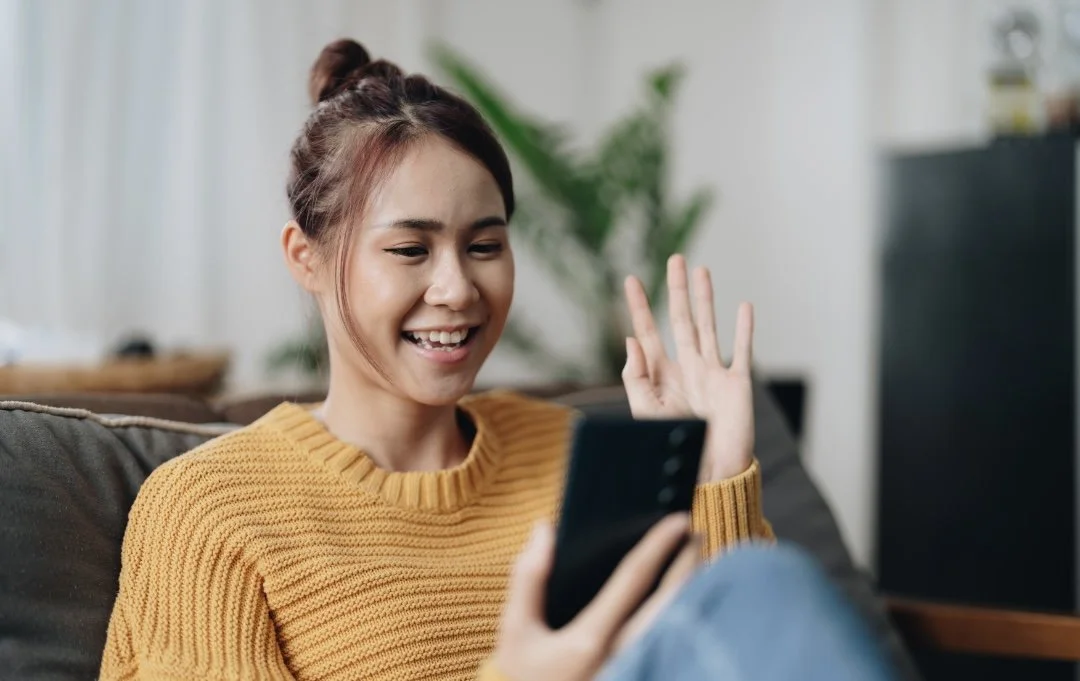 A young woman with brown hair in a bun, wearing a mustard yellow sweater, sitting on a couch, smiling, and waving at her phone screen.